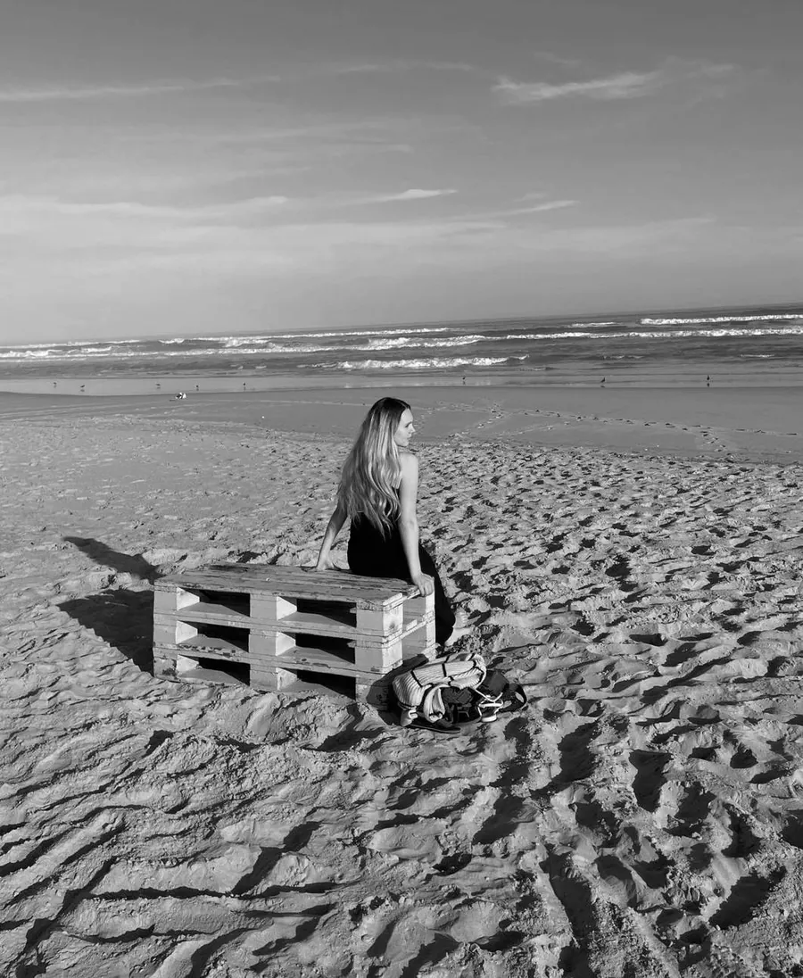 Merdene Coetzee, designer and founder of Merdene Design Studio, seated on a wooden pallet on a beach